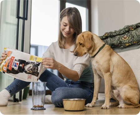 Ženska naliva hrano znamke Hill's Pet Nutrition v merilni lonček, medtem ko pes pozorno opazuje.